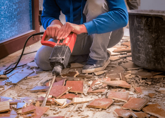Floor removal service Houston specialist using a demolition hammer to break up large tiles into a debris bucket.